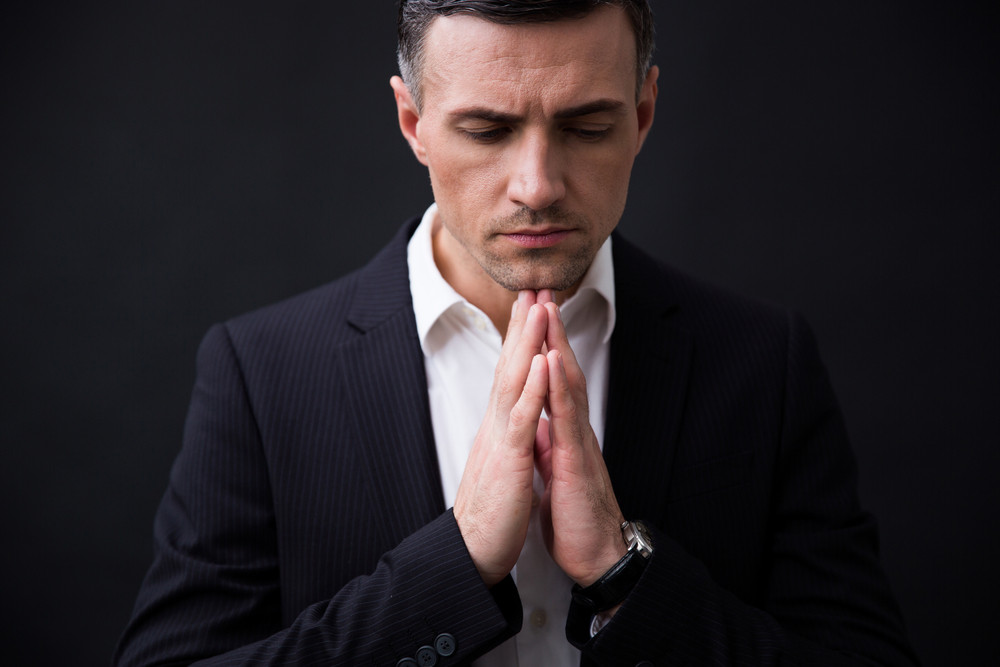 Portrait of a businessman in formal wear praying over black background 